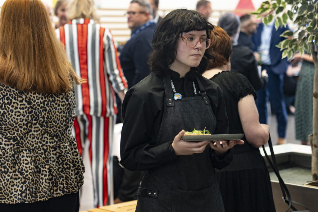 A student wearing a black apron with a "STUDENT" lanyard, holds a small plate with food, looking down. Other people are blurred in the background.
