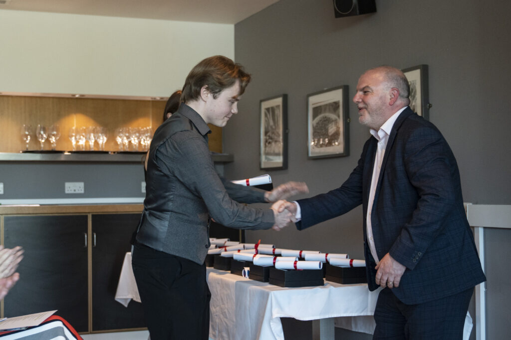 A person in a dark shirt shakes hands with a smiling bald man in a suit, both holding rolled-up certificates, over a table laden with more certificates.
