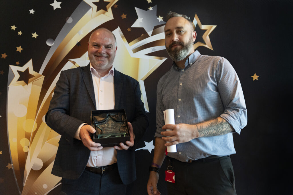  A smiling man in a suit holding an award and a smiling man in a light shirt holding a certificate, against a star-patterned background.