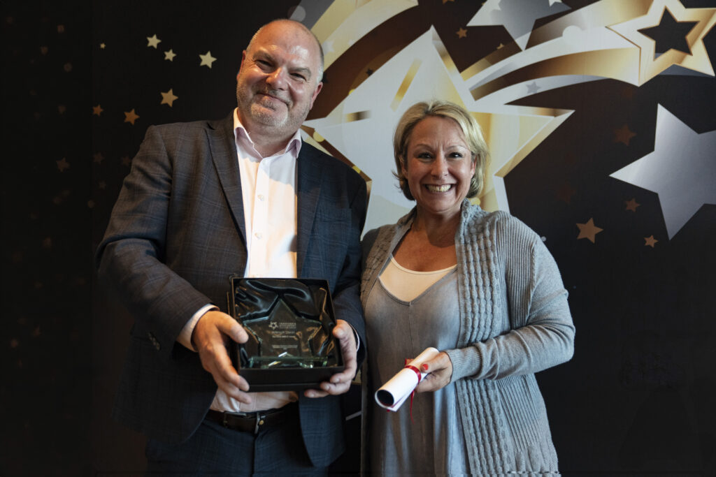 A smiling man in a suit holds a boxed award, standing next to a smiling woman with short blonde hair in a grey cardigan, who holds a rolled-up certificate. They are against a star-patterned background.